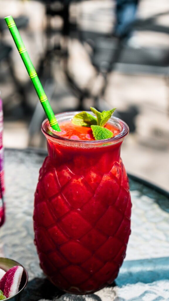 Un verre original en forme d’ananas rempli d’un cocktail rouge vibrant, garni de feuilles de menthe fraîche et accompagné d’une paille verte.