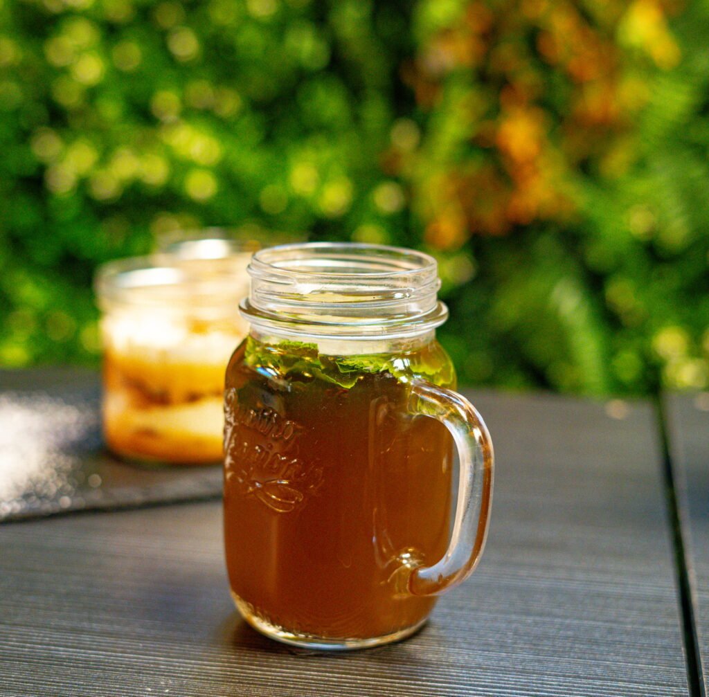 Une tasse en verre contenant un thé chaud avec des feuilles de menthe, posée sur une table en extérieur, avec un arrière-plan verdoyant.