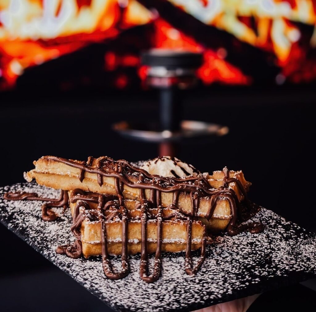  Assiette gourmande de gaufres croustillantes nappées de chocolat, accompagnée d'une boule de glace vanille et saupoudrée de sucre glace, avec un narguilé en arrière-plan.