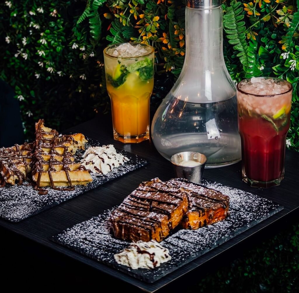 Assortiment de desserts gourmands avec une chicha moderne et des cocktails colorés, disposés sur une table dans un cadre végétal élégant.